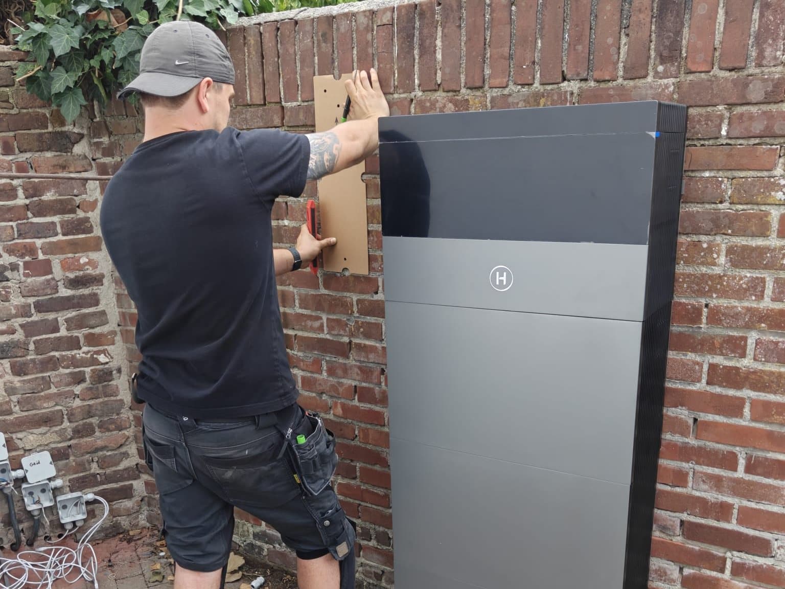 Monteur met pet, van achteren gezien, houdt een kartonnen mal tegen een bakstenen muur naast een grijze thuisbatterij.