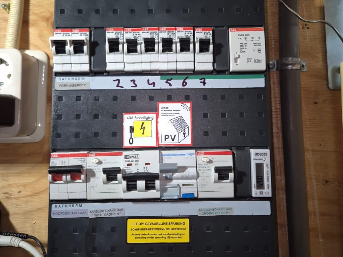 Groepenkast met een rij installatieautomaten, een sticker voor de zonnepanelen en een gele waarschuwingssticker.