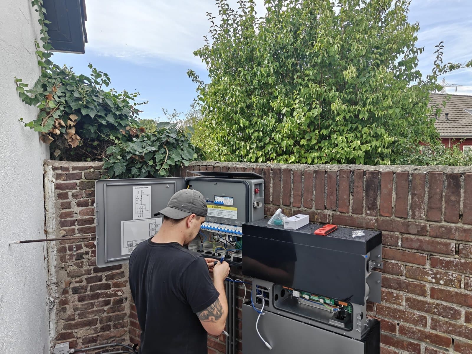 Monteur met pet werkt aan een geopende grijze kast aan een bakstenen tuinmuur, met daarnaast de thuisbatterij waarvan de bovenkant nog open is.