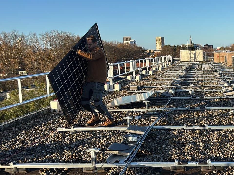 Monteur in een bruine trui draagt een zwart zonnepaneel over een plat grinddak vol montagerails, met daarachter een stad.