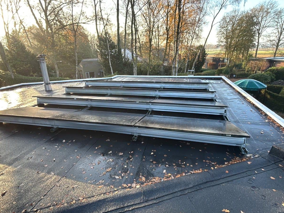 Rijen zwarte zonnepanelen op schuine aluminium frames op een nat plat dak, met bomen in laag zonlicht.