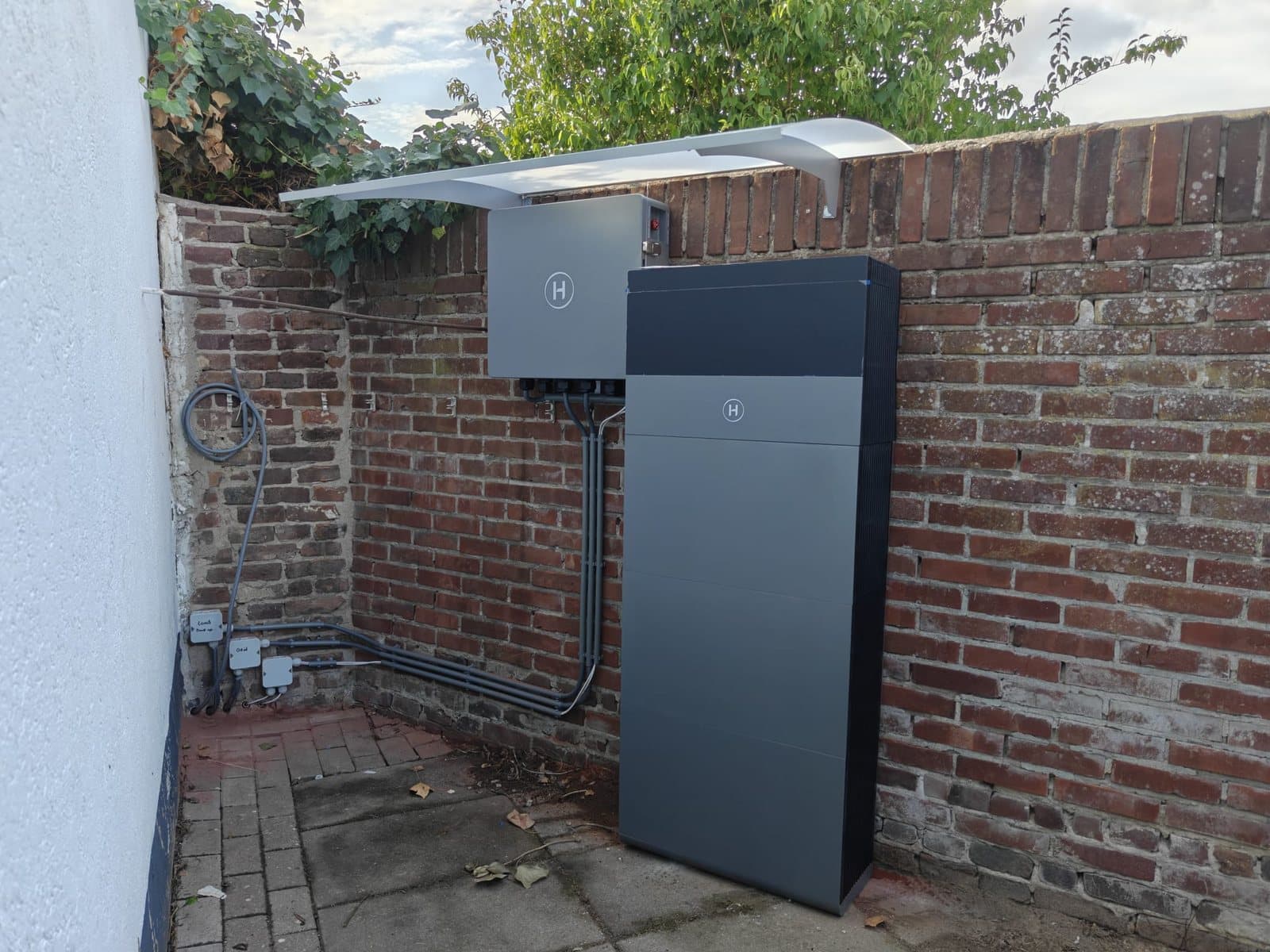 Grijze thuisbatterij en een kleinere grijze kast met het logo van Hoymiles tegen een bakstenen tuinmuur, met de kabels in grijze buizen langs de muur.